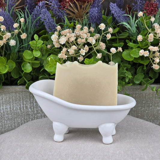 A bar of Jarhead Aromatics Eucalyptus + Mint Handmade Natural Goat Milk Artisan Soap rests in a small white ceramic bathtub dish, with green leaves and purple and white flowers in the background.