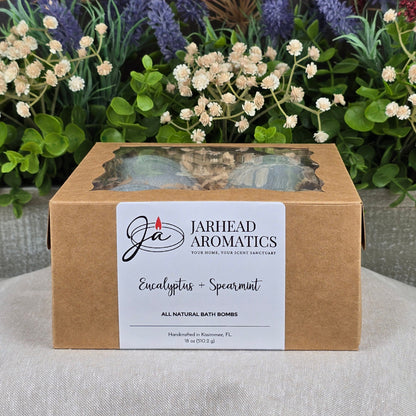 A brown box labeled Jarhead Aromatics Eucalyptus + Spearmint Bath Bomb - All Natural rests on a gray surface, with assorted artificial flowers and green leaves in the background.