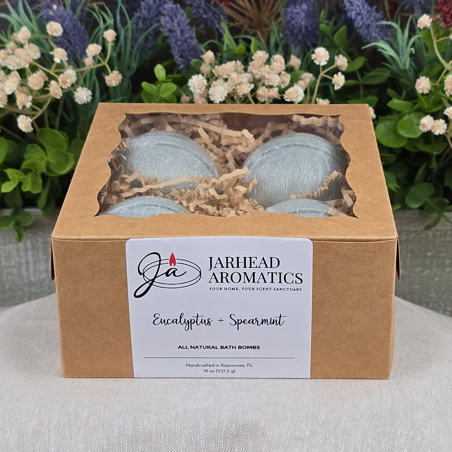 A box of Jarhead Aromatics Eucalyptus + Spearmint Bath Bomb - All Natural sits on a gray surface, with a labeled front and windowed top, surrounded by artificial greenery and purple flowers in the background.