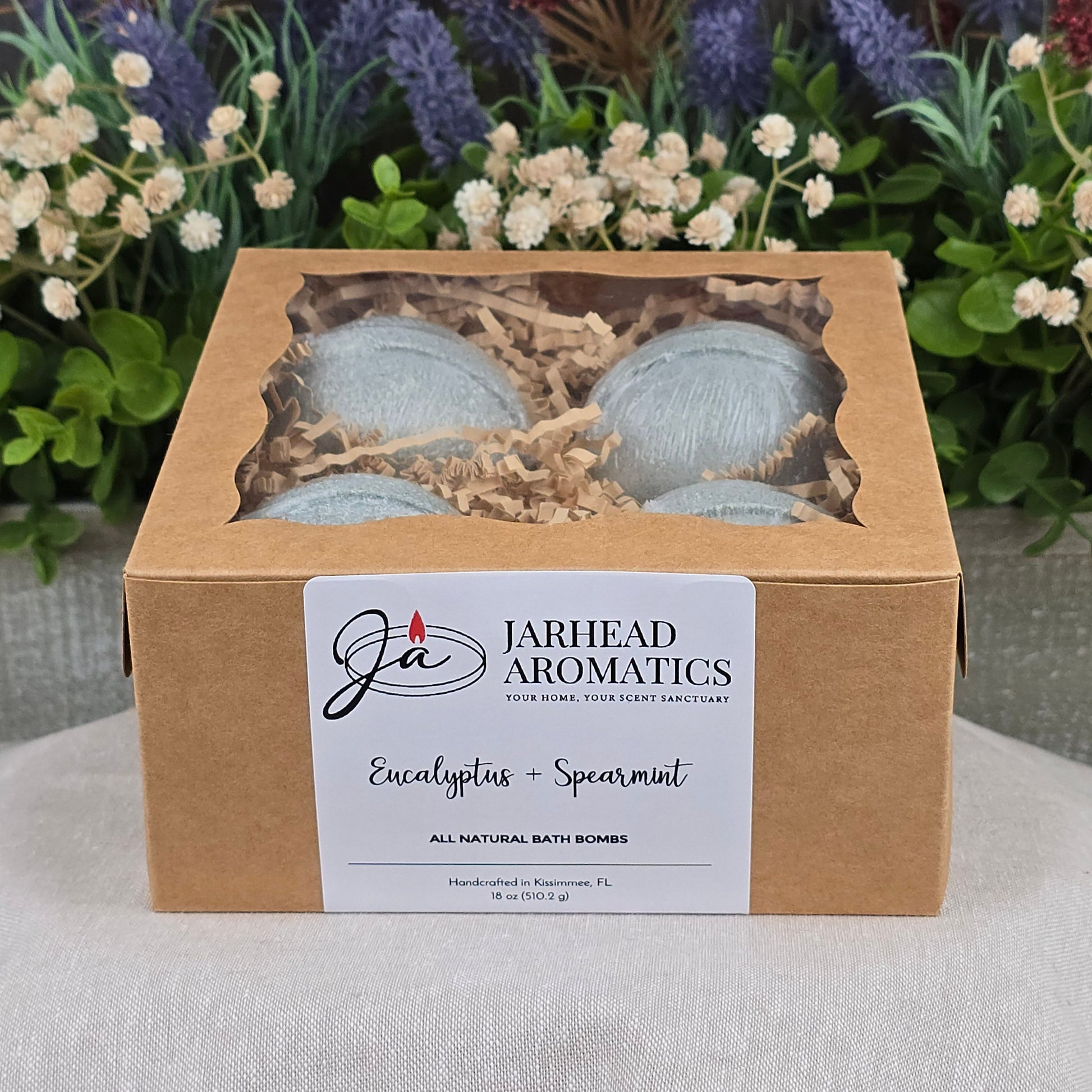 A box of Jarhead Aromatics Eucalyptus + Spearmint Bath Bomb - All Natural sits on a gray surface, with a labeled front and windowed top, surrounded by artificial greenery and purple flowers in the background.