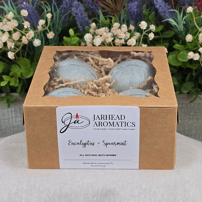 A box of Jarhead Aromatics Eucalyptus + Spearmint Bath Bomb - All Natural sits on a gray surface, with a labeled front and windowed top, surrounded by artificial greenery and purple flowers in the background.