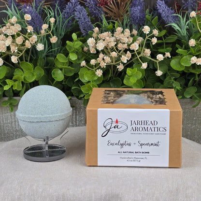 A handmade Eucalyptus + Spearmint Bath Bomb by Jarhead Aromatics sits on a stand beside its brown box, with colorful artificial flowers in the background.