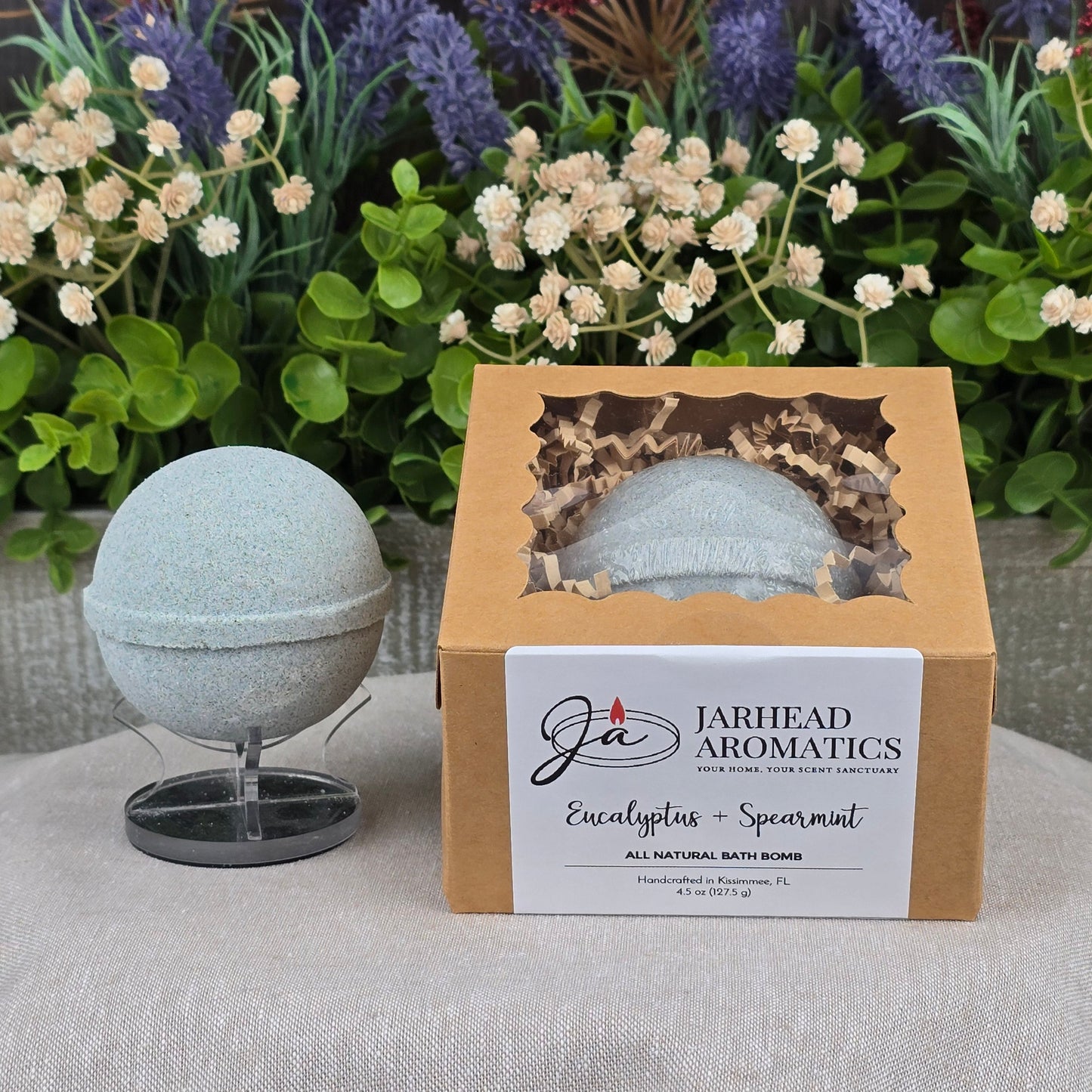 A light green, handcrafted Eucalyptus + Spearmint Bath Bomb by Jarhead Aromatics sits on a stand beside its box. Decorative greenery and flowers are in the background on a gray surface.