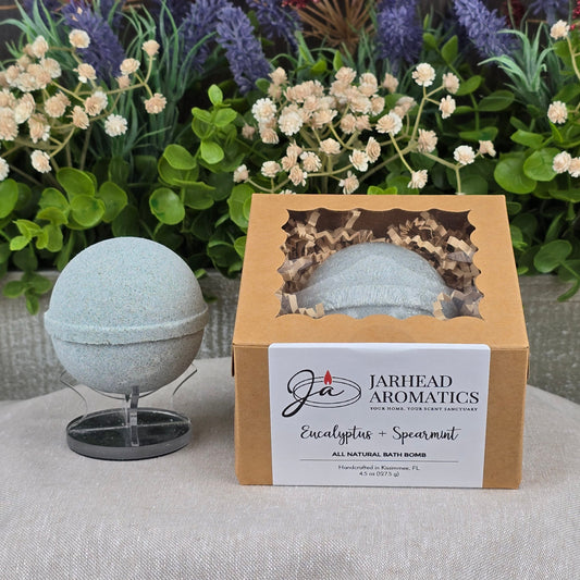 A light green, handcrafted Eucalyptus + Spearmint Bath Bomb by Jarhead Aromatics sits on a stand beside its box. Decorative greenery and flowers are in the background on a gray surface.