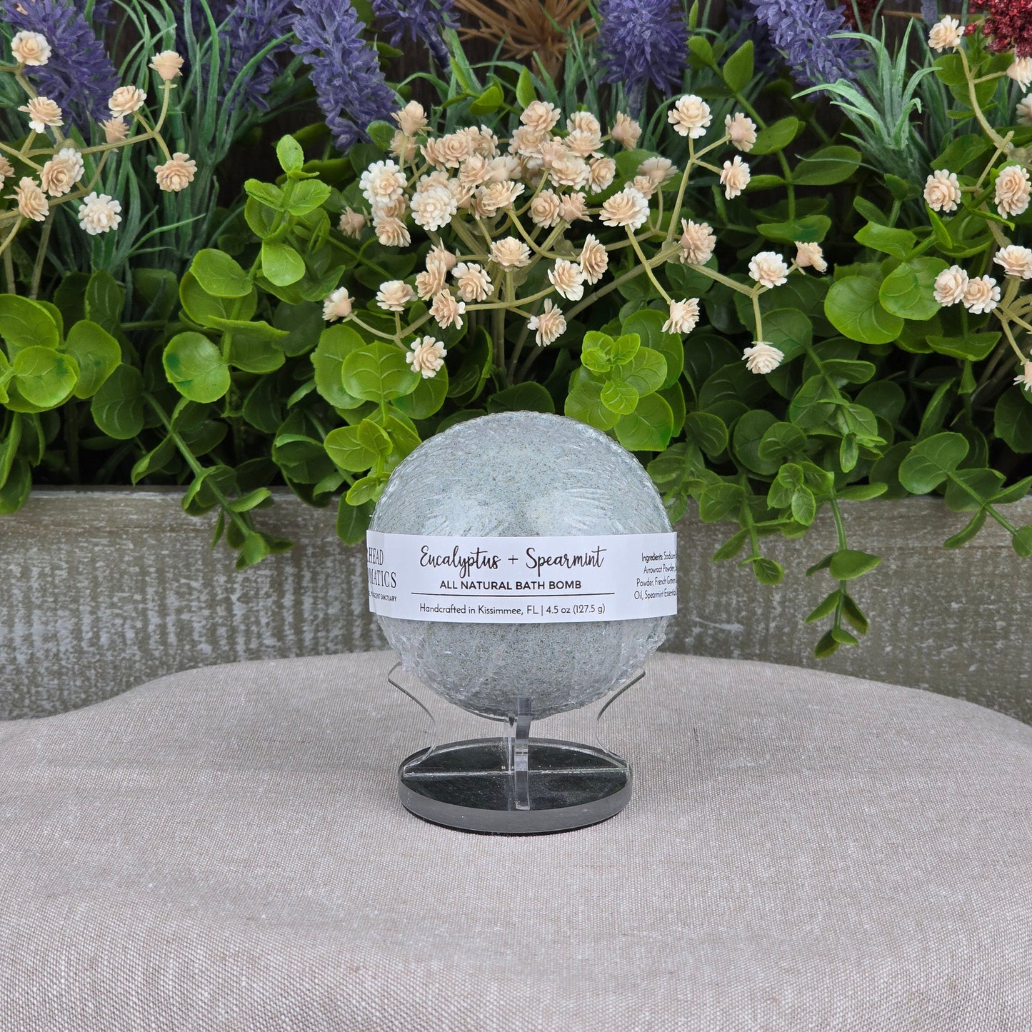 The Eucalyptus + Spearmint Bath Bomb - All Natural by Jarhead Aromatics, in light green, is displayed on a stand surrounded by artificial greenery and purple and red flowers against a gray background.