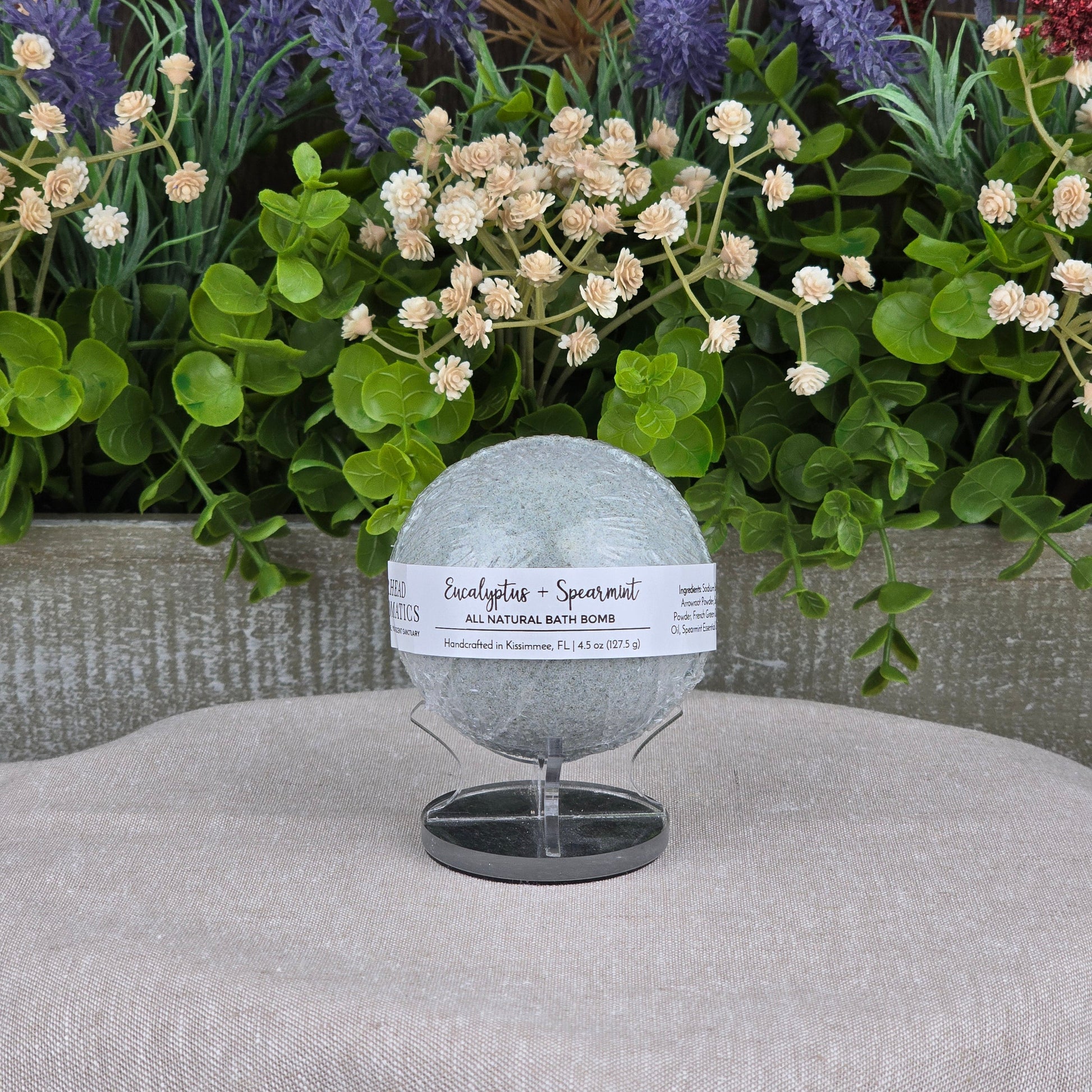 The Eucalyptus + Spearmint Bath Bomb - All Natural by Jarhead Aromatics, in light green, is displayed on a stand surrounded by artificial greenery and purple and red flowers against a gray background.