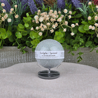 The Eucalyptus + Spearmint Bath Bomb - All Natural by Jarhead Aromatics, in light green, is displayed on a stand surrounded by artificial greenery and purple and red flowers against a gray background.