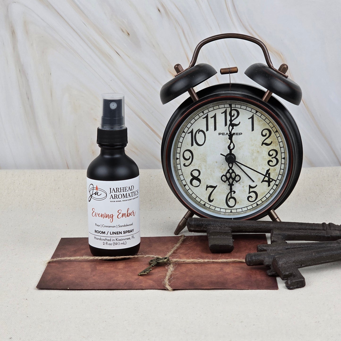 A vintage alarm clock, three antique keys, a tied brown envelope, and Jarhead Aromatics Evening Ember Room & Linen Spray—Plum, Smoke & Sandalwood—are displayed on a light surface with a marble background.