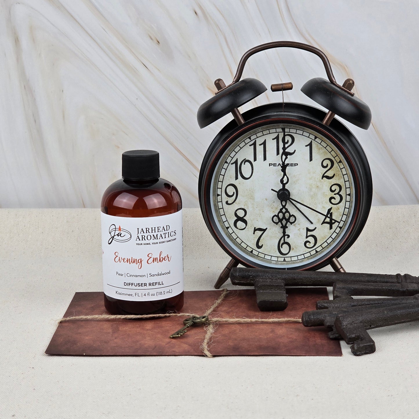 A bottle of Jarhead Aromatics Evening Ember Diffuser Refill | Plum, Sandalwood & Smoke sits beside a vintage black alarm clock and antique keys atop a leather-bound book, perfect for cozy home fragrance in any space.