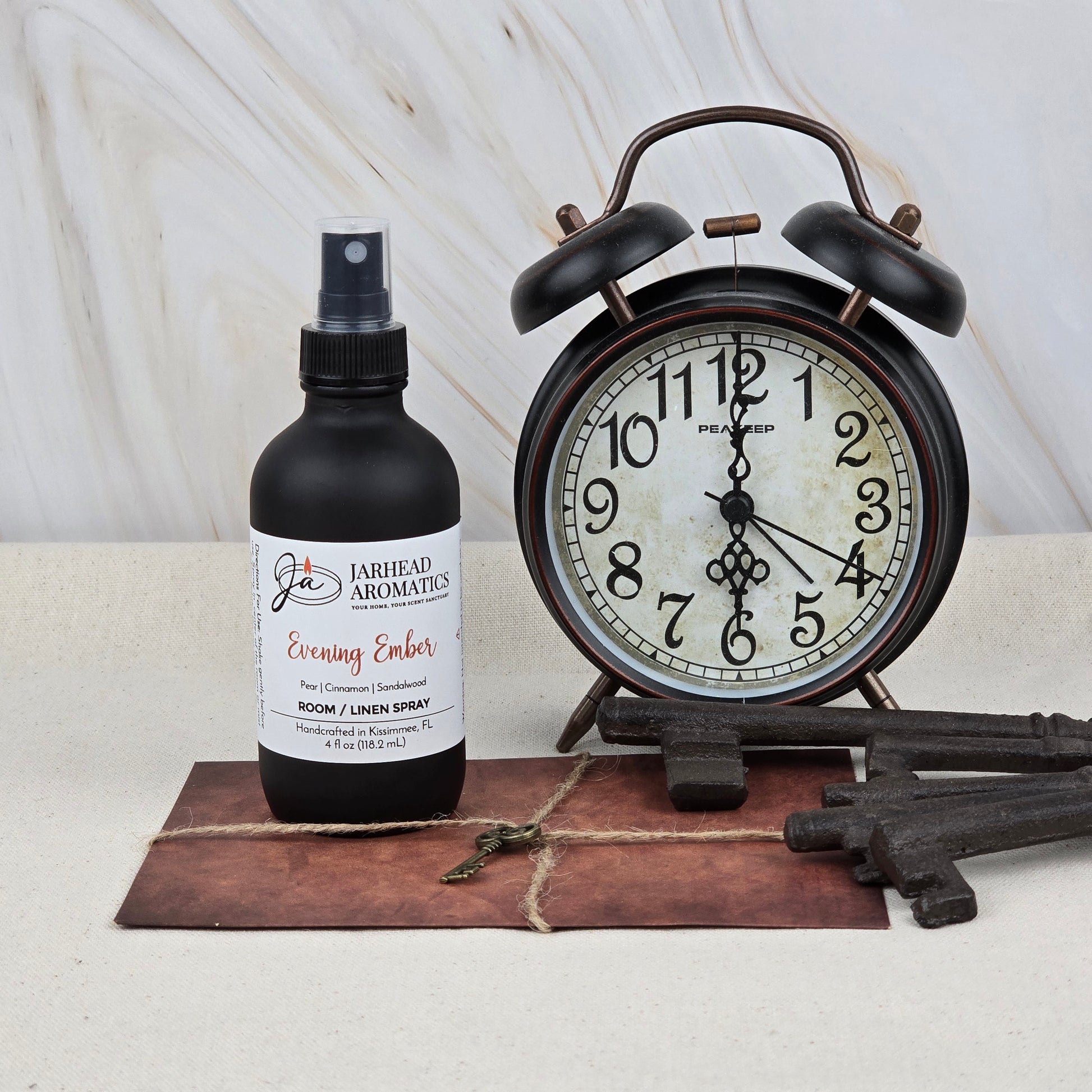 A Jarhead Aromatics Evening Ember Room & Linen Spray (Plum, Smoke & Sandalwood) sits beside a vintage black alarm clock, three old-fashioned keys, and a brown envelope tied with string.