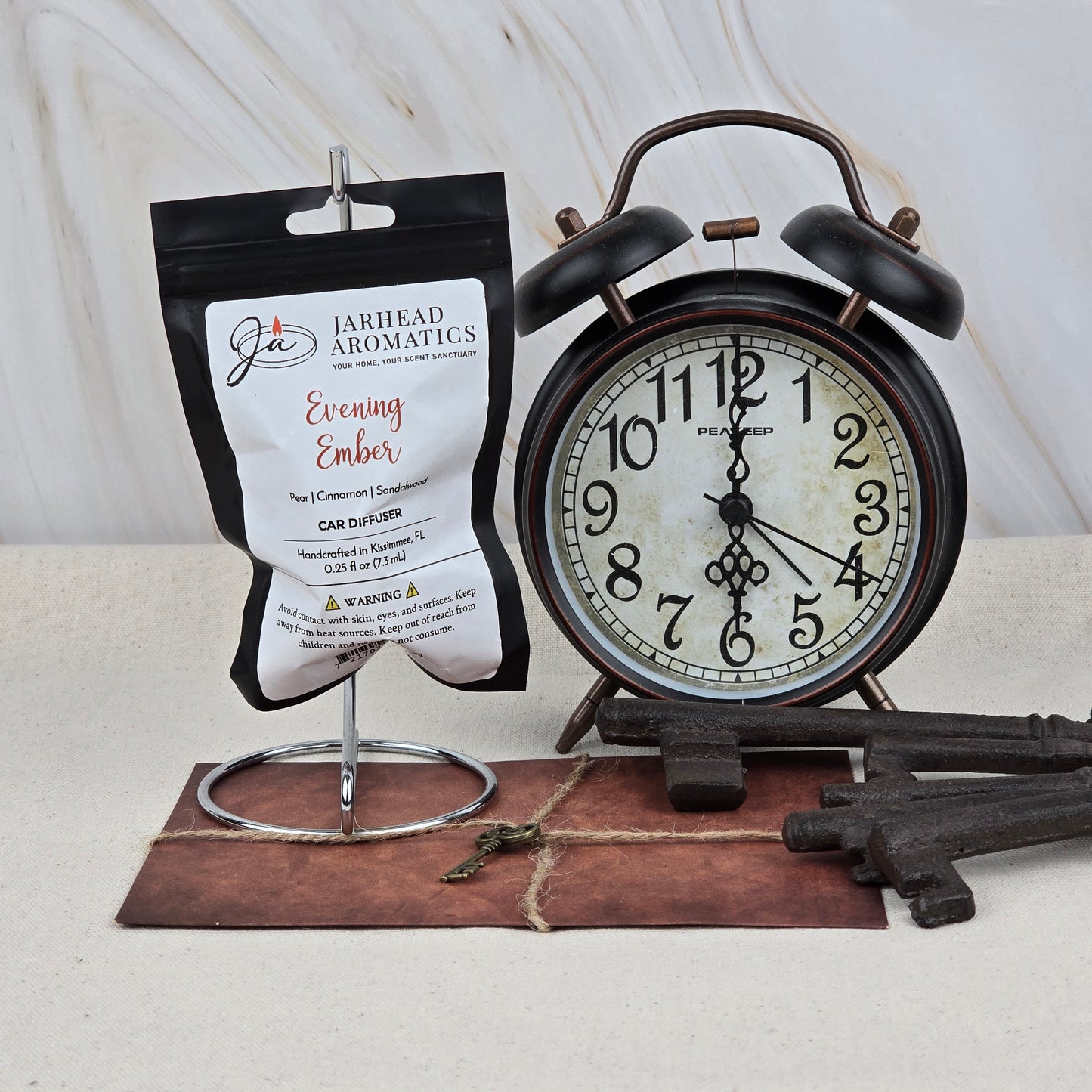 A Jarhead Aromatics Evening Ember Car Diffuser | Plum, Sandalwood & Smoke Car Air Freshener sits on a metal stand next to a vintage black alarm clock, three antique keys, and an old key atop a brown envelope, all on a light surface.