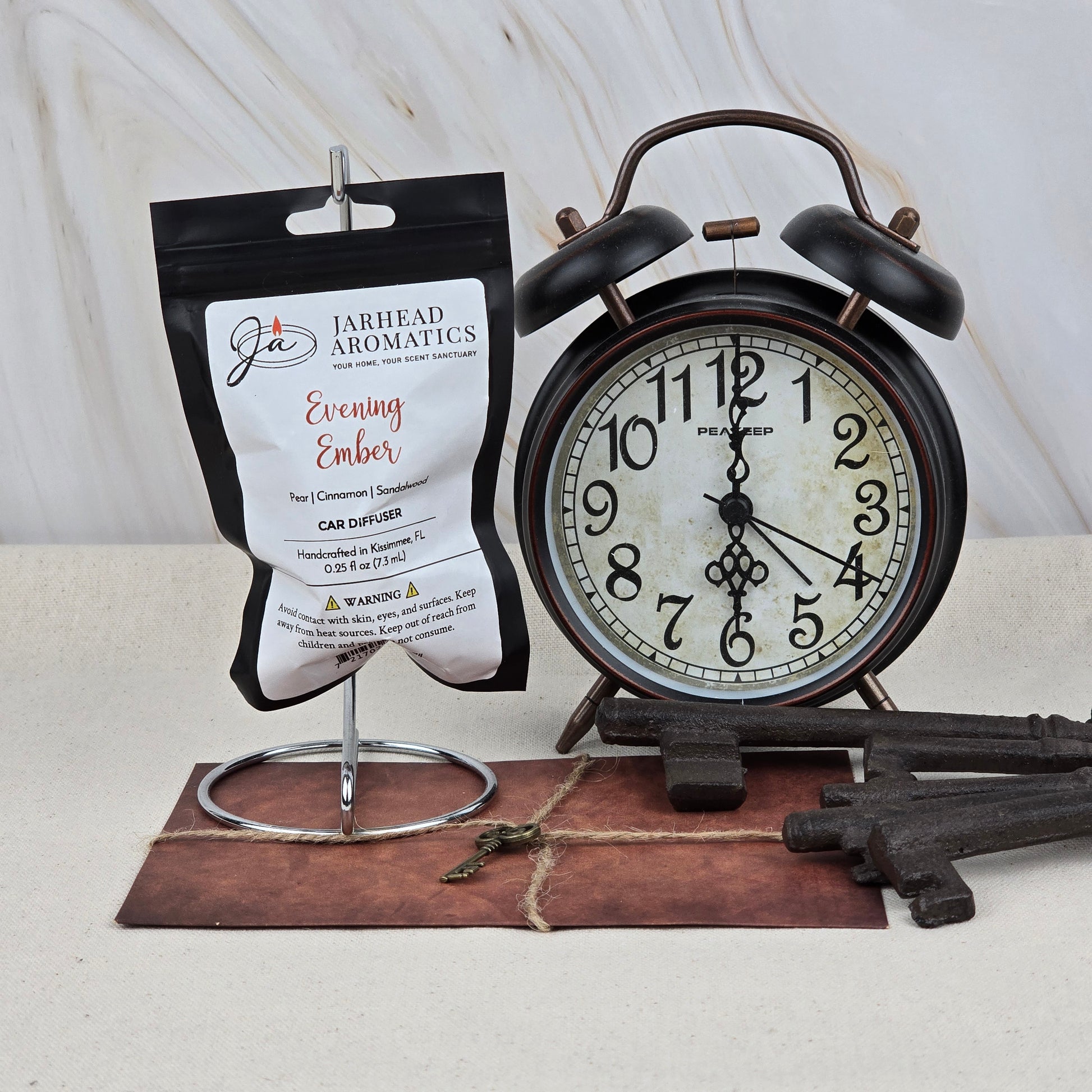 A Jarhead Aromatics Evening Ember Car Diffuser | Plum, Sandalwood & Smoke Car Air Freshener sits on a metal stand next to a vintage black alarm clock, three antique keys, and an old key atop a brown envelope, all on a light surface.