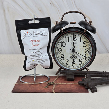 A Jarhead Aromatics Evening Ember Car Diffuser | Plum, Sandalwood & Smoke Car Air Freshener sits on a metal stand next to a vintage black alarm clock, three antique keys, and an old key atop a brown envelope, all on a light surface.