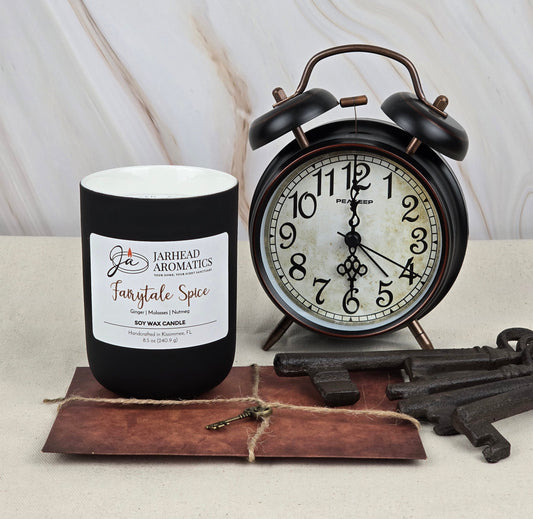 A Jarhead Aromatics black jar candle, Fairytale Spice - Soy Wax Candle, emits a gingerbread scent beside a vintage alarm clock. Nearby, antique keys rest on vintage paper atop a beige surface with a marble backdrop.