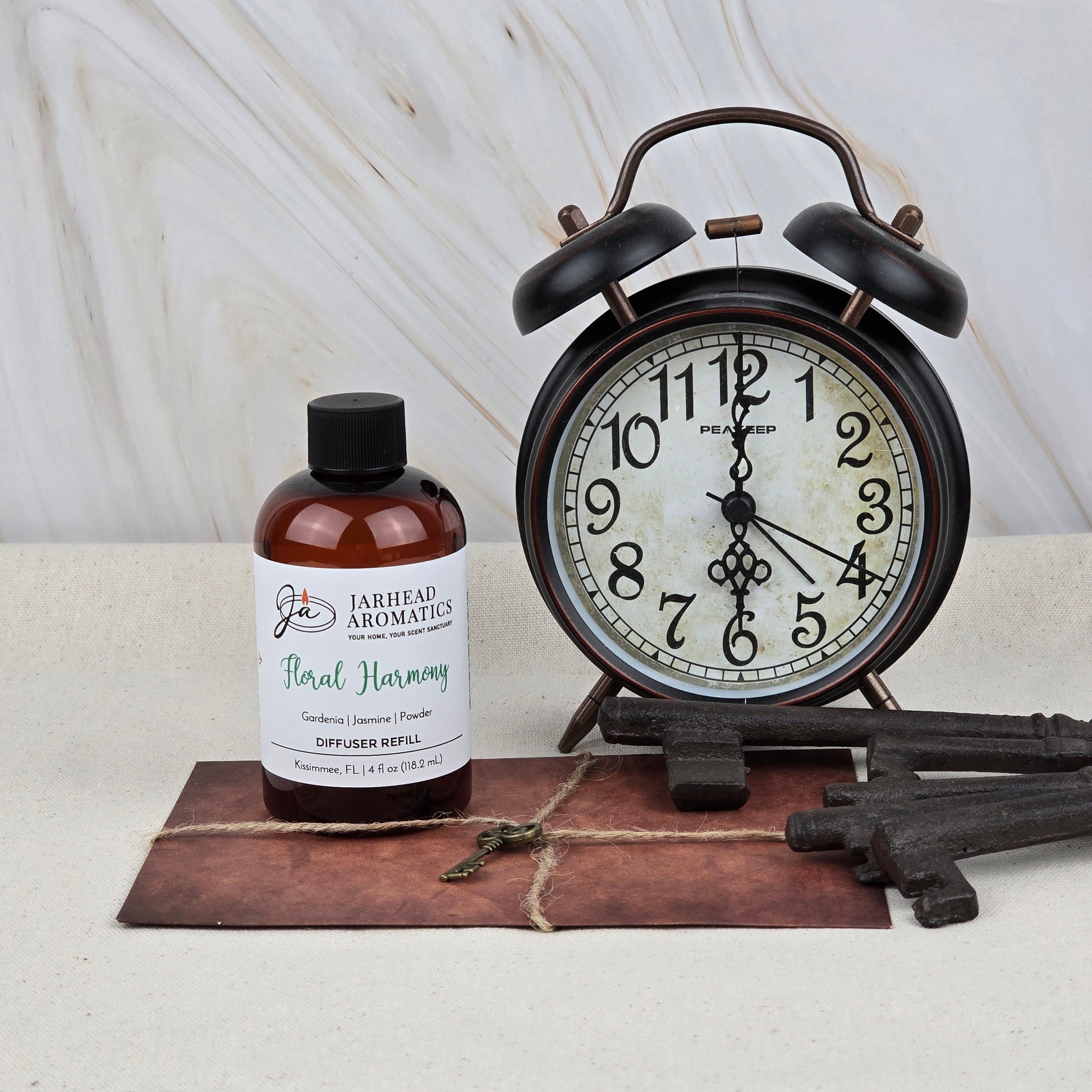 A bottle of Jarhead Aromatics Floral Harmony Diffuser Refill—Gardenia, Jasmine & Tuberose—rests beside a vintage black alarm clock, two large antique keys, and a small brass key on brown leather. Marble background.