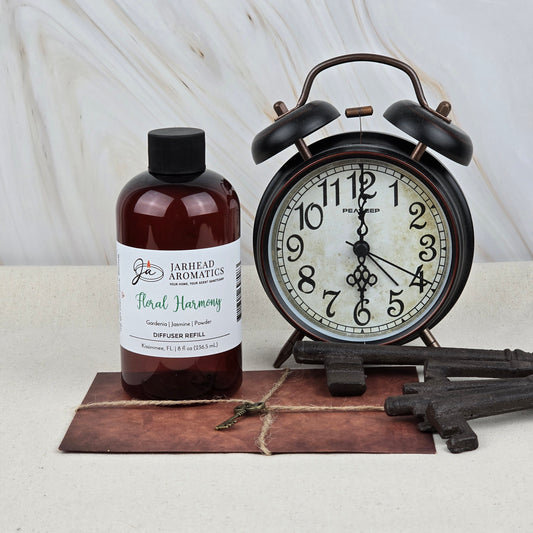 A bottle of Jarhead Aromatics Floral Harmony Diffuser Refill | Gardenia, Jasmine & Tuberose Home Fragrance Oil Refill stands next to a vintage alarm clock and keys on a rustic brown surface with a light marble background.