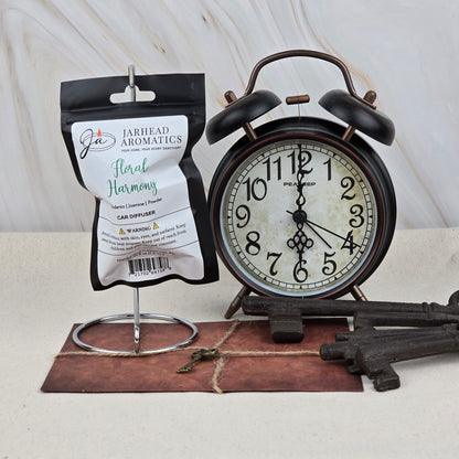 A Jarhead Aromatics Floral Harmony Car Diffuser (Gardenia, Jasmine & Tuberose) hangs by a stand near a vintage black alarm clock, two large antique keys, a small key, and a brown envelope on a light surface.