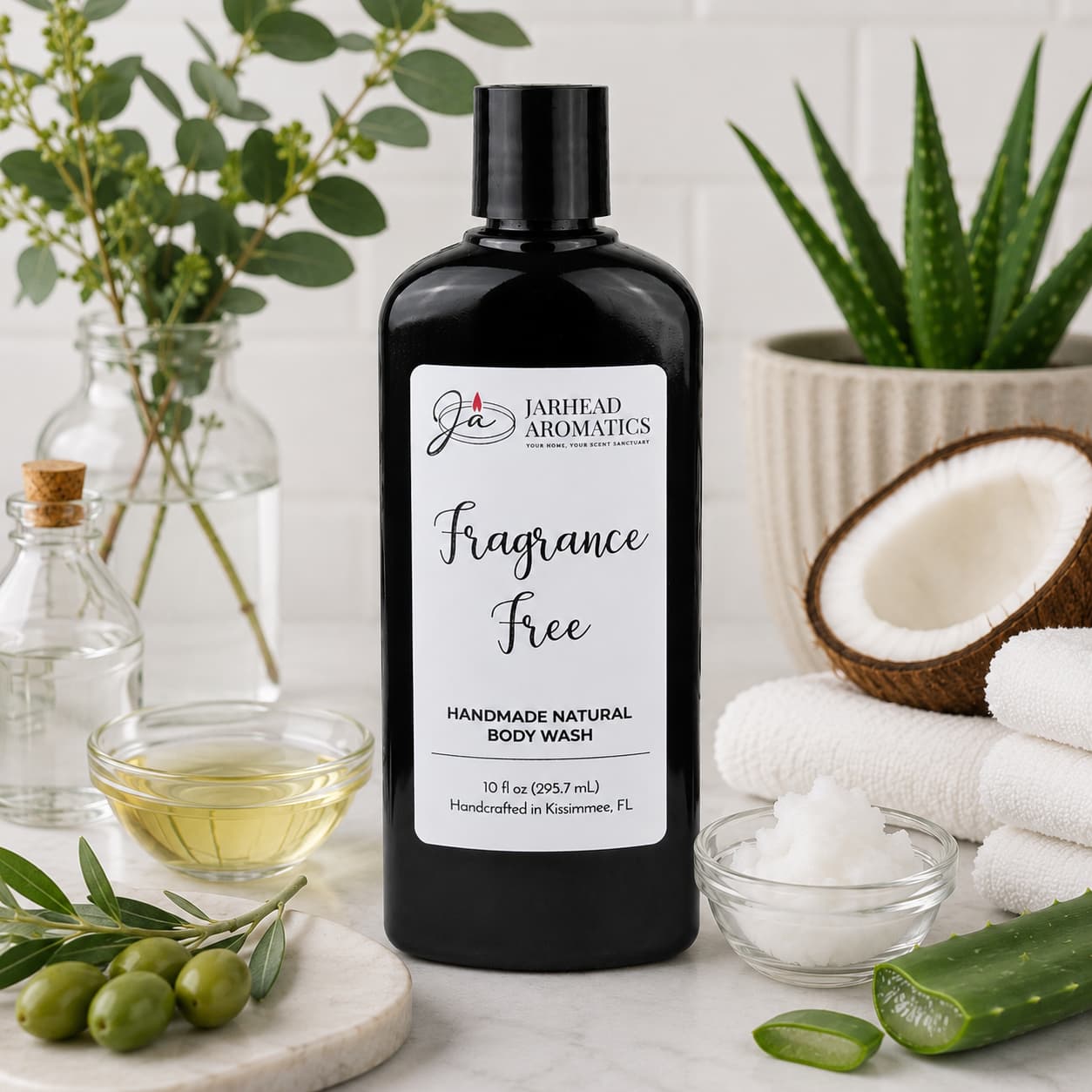 A black bottle of Jarhead Aromatics Fragrance-Free Handmade Natural Body Wash, featuring organic aloe and plant-based oils, is displayed on a white countertop surrounded by olives, coconut, oil, towels, greenery, and a small bowl of white cream. This gentle unscented body wash offers pure and soothing cleansing with its nourishing ingredients.