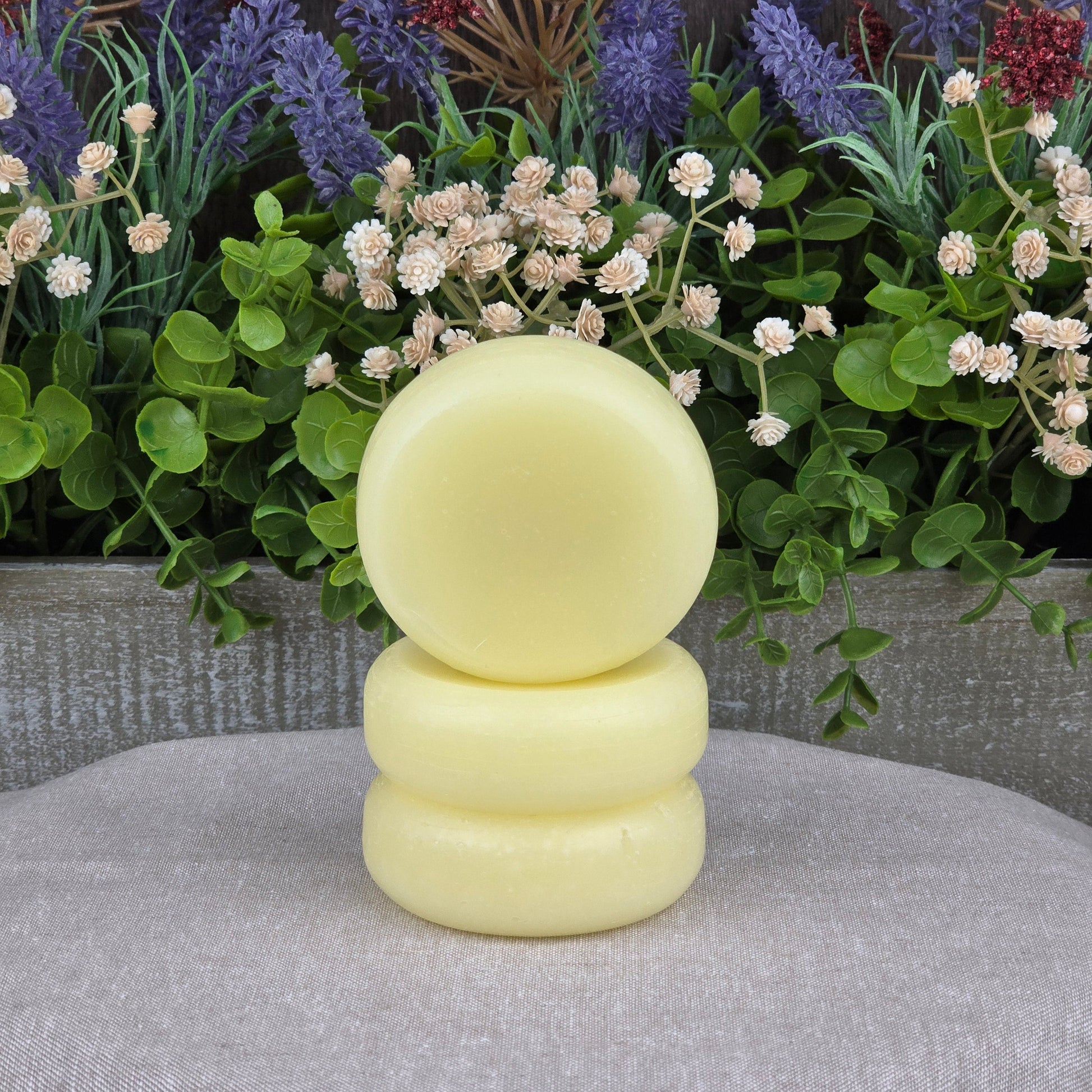 Three round, pale yellow Jarhead Aromatics Fragrance-Free Handmade Conditioner Bars are stacked before green leaves and colorful artificial flowers on a light gray surface—ideal for anyone seeking zero-waste, all-natural hair care.