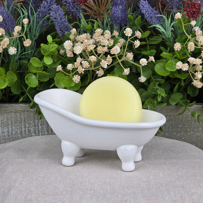 A Fragrance-Free Handmade Conditioner Bar from Jarhead Aromatics rests in a white clawfoot tub dish, with greenery and purple flowers in the background—an all-natural, zero-waste hair care solution.