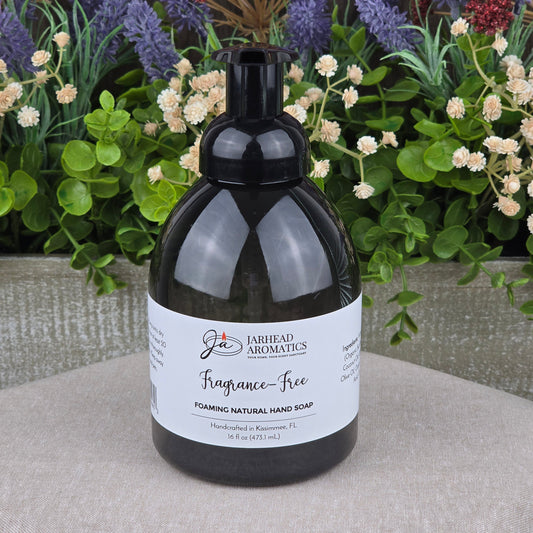 A black pump bottle of Jarhead Aromatics Fragrance-Free Foaming Natural Hand Soap rests on a gray surface, with assorted green foliage and purple flowers in the background.