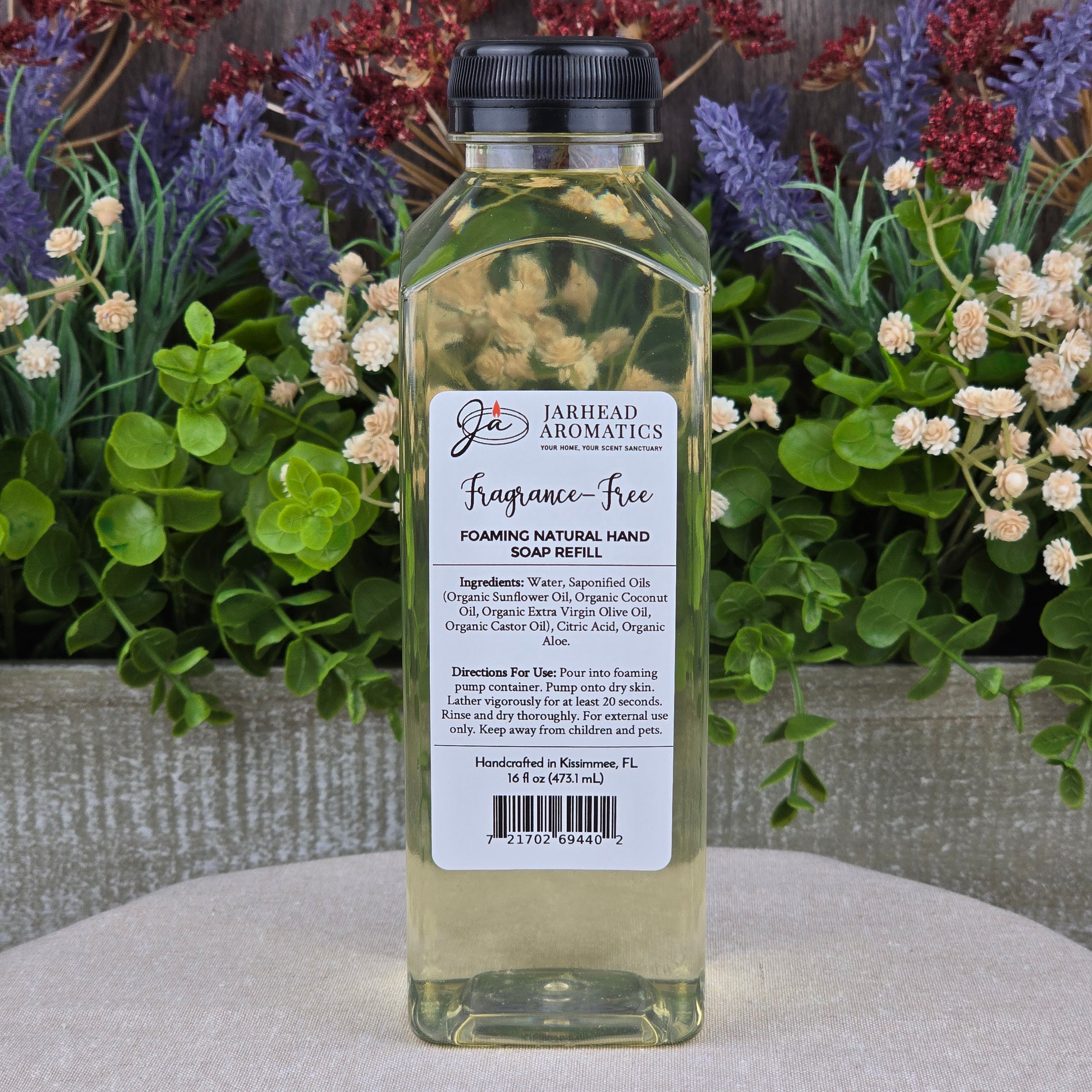 A clear plastic bottle of Jarhead Aromatics Fragrance-Free Foaming Natural Hand Soap, suitable for sensitive skin, stands on a gray surface with colorful artificial flowers behind it. The label lists ingredients and directions.