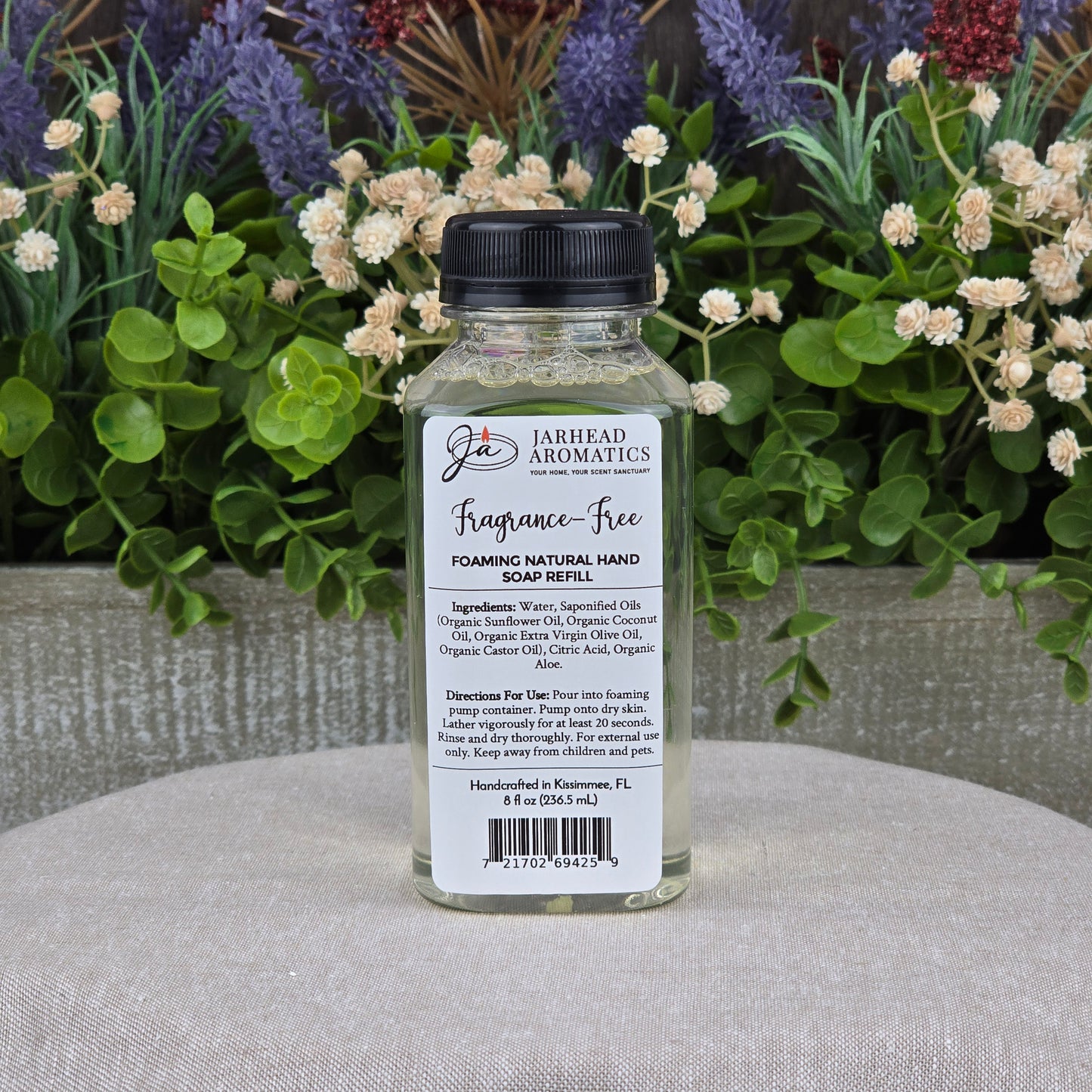 A clear bottle of Jarhead Aromatics Fragrance-Free Foaming Natural Hand Soap sits on a light gray surface with green plants and purple flowers. Ideal for sensitive skin, it includes ingredients and directions for use on the label.