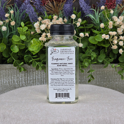 A clear bottle of Jarhead Aromatics Fragrance-Free Foaming Natural Hand Soap sits on a light gray surface with green plants and purple flowers. Ideal for sensitive skin, it includes ingredients and directions for use on the label.