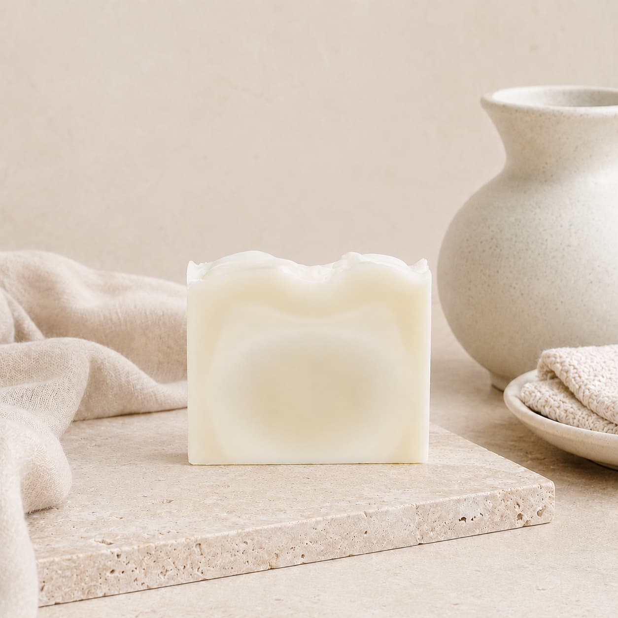 A rectangular bar of Fragrance-Free Handmade Natural Tallow Artisan Soap from Jarhead Aromatics rests on a beige stone surface, accompanied by a soft towel, a ceramic vase, and a dish in the background to evoke a minimalist, spa-like atmosphere.