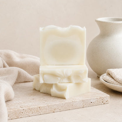 Three bars of Jarhead Aromatics Fragrance-Free Handmade Natural Tallow Artisan Soap are stacked on a stone surface, accompanied by a beige cloth, a white ceramic vase, and a small plate with textured fabric. The scene features a neutral, minimalistic background that highlights the unscented, minimal-ingredient tallow soap.