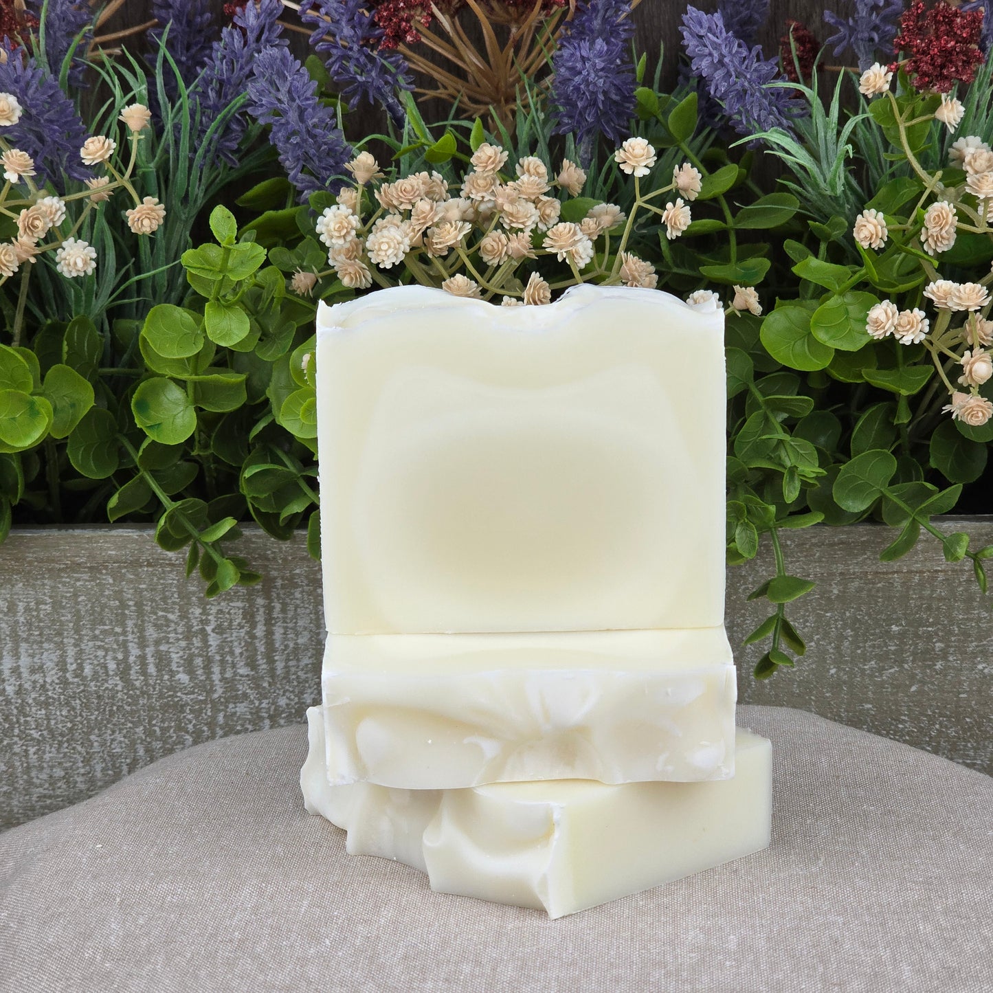 Three handmade Fragrance-Free Natural Tallow Artisan Soaps by Jarhead Aromatics are stacked on a beige cloth, surrounded by flowers and greenery, highlighting their gentle, unscented formula for sensitive skin.