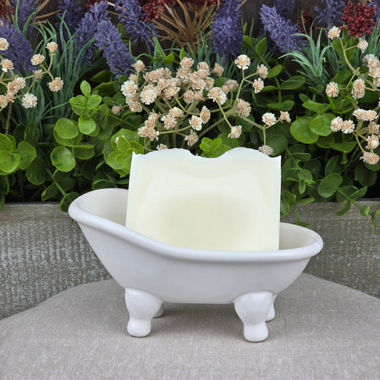 A bar of Jarhead Aromatics Fragrance-Free Handmade Natural Tallow Artisan Soap for sensitive skin rests in a white bathtub-shaped soap dish, set before a backdrop of green and purple artificial flowers.