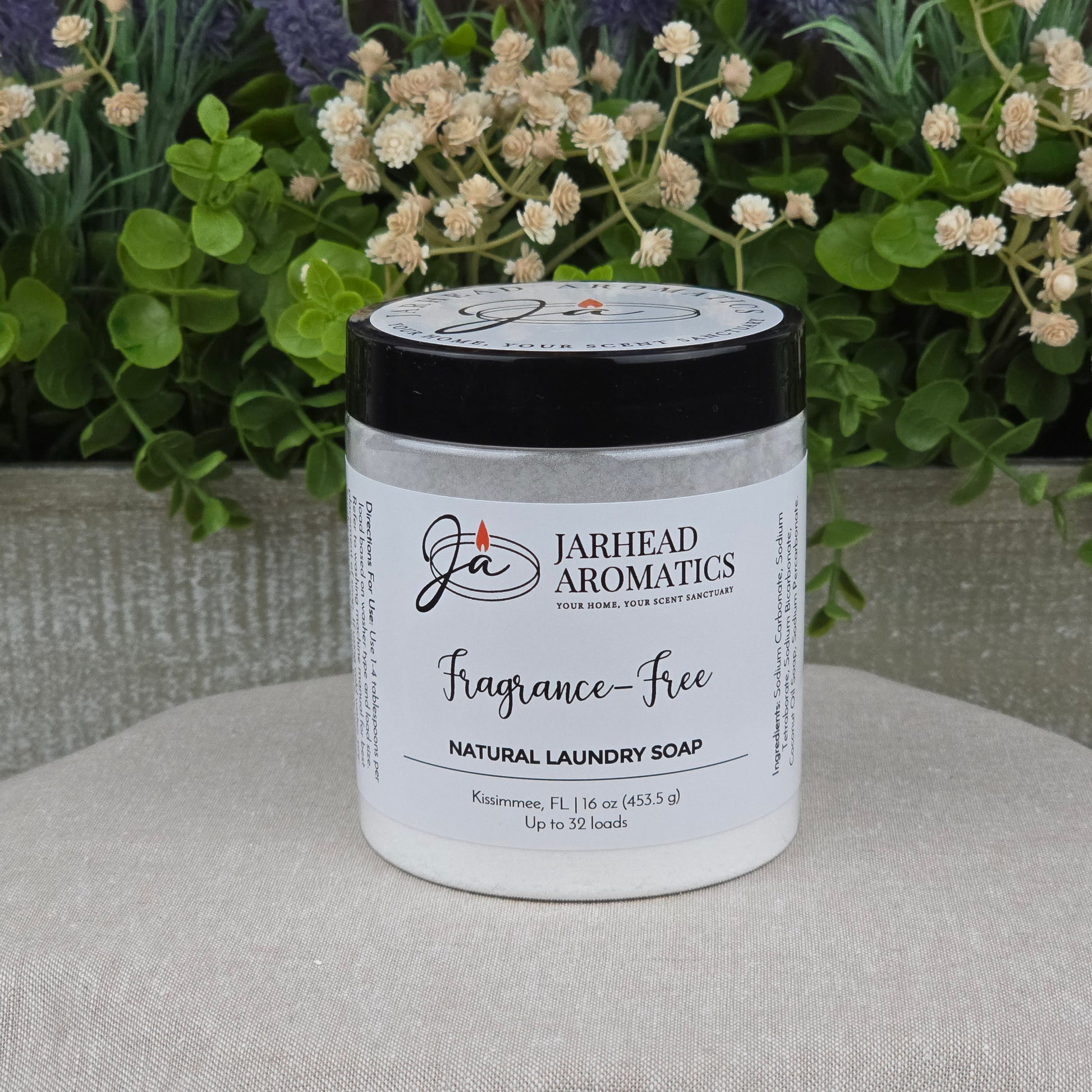 A jar of Jarhead Aromatics Fragrance-Free Handmade Natural Laundry Soap, a non-toxic powder detergent for sensitive skin, rests on a light surface with green plants and flowers in the background. The white jar has a black lid and clean label.