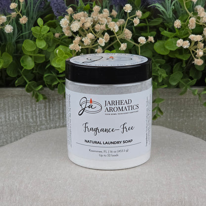 A jar of Jarhead Aromatics Fragrance-Free Handmade Natural Laundry Soap, a non-toxic powder detergent for sensitive skin, rests on a light surface with green plants and flowers in the background. The white jar has a black lid and clean label.