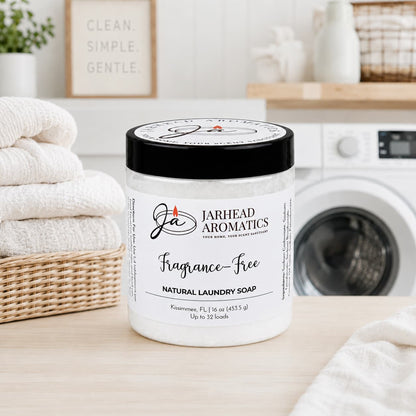 A container of Jarhead Aromatics Fragrance-Free Handmade Natural Laundry Soap sits on a table in a laundry room alongside folded towels, a plant, and a front-loading washing machine in the background. This unscented powdered laundry detergent offers gentle, effective cleaning without added fragrance—ideal for those who prefer pure, natural care.