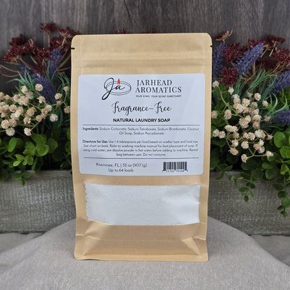A brown bag of Jarhead Aromatics Fragrance-Free Handmade Natural Laundry Soap, a non-toxic powder detergent for sensitive skin, stands on a gray surface with its label visible; flowers and greenery decorate the background.
