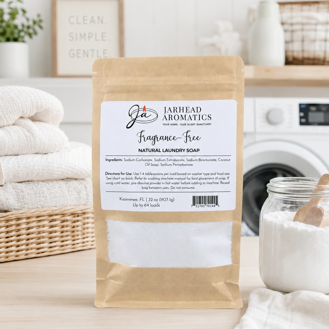 A brown paper bag labeled "Fragrance-Free Handmade Natural Laundry Soap | Unscented Powdered Laundry Detergent" from Jarhead Aromatics—a handmade, fragrance-free laundry soap—rests on a laundry room countertop, surrounded by stacked towels, a washing machine, and a jar of powder in the background.