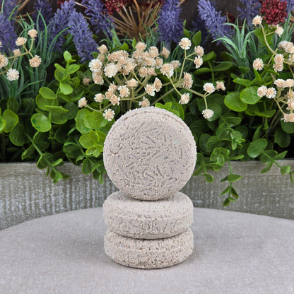 Three round, textured gray Fragrance-Free Handmade Natural Shampoo & Conditioner Bars by Jarhead Aromatics are stacked on a gray fabric before vibrant artificial flowers and green leaves, offering a handmade, natural alternative for hair care.