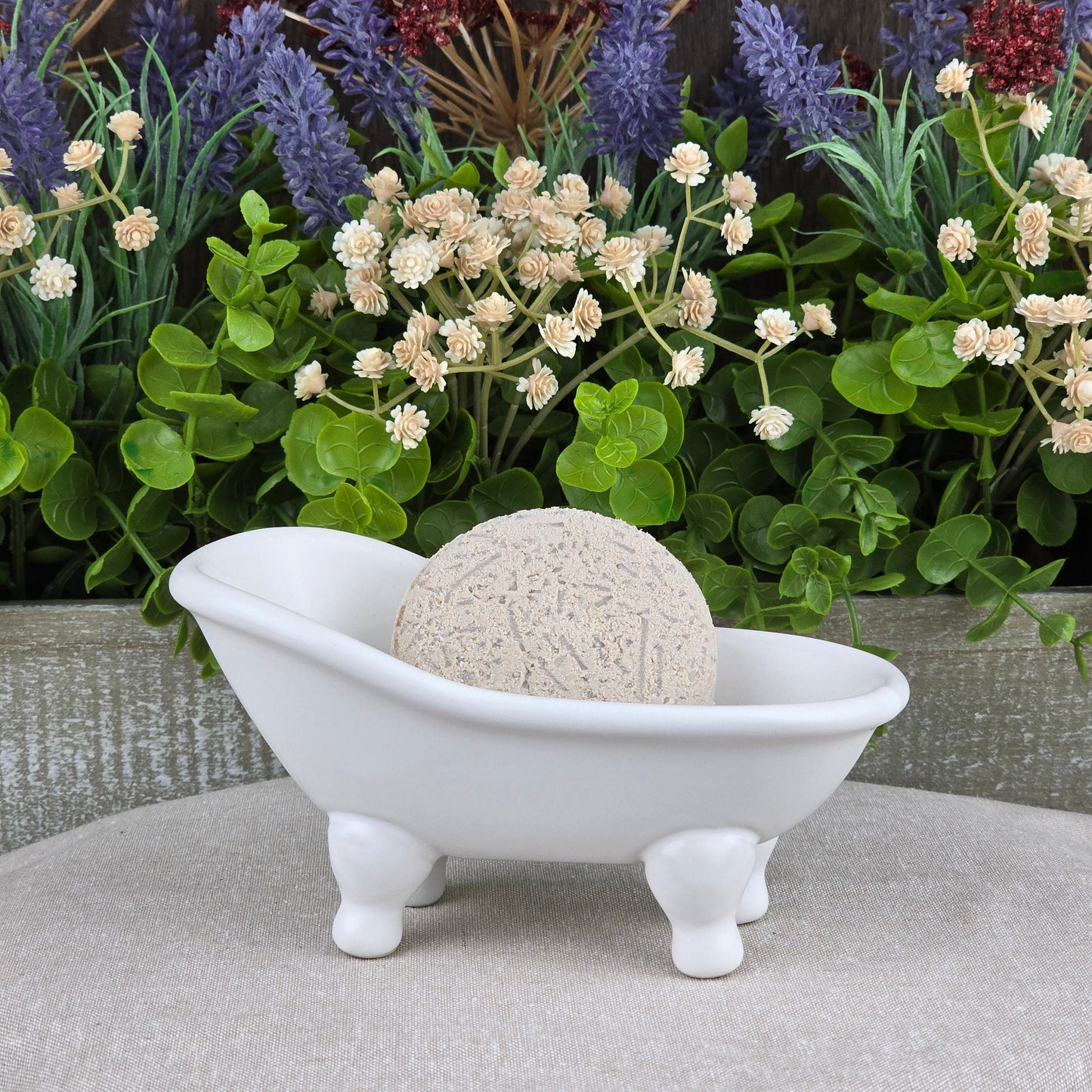 The Jarhead Aromatics Fragrance-Free Handmade Natural Shampoo & Conditioner Bar, beige and round, rests in a white clawfoot dish on a neutral cloth, with green leaves and colorful faux lavender and burgundy flowers behind it.