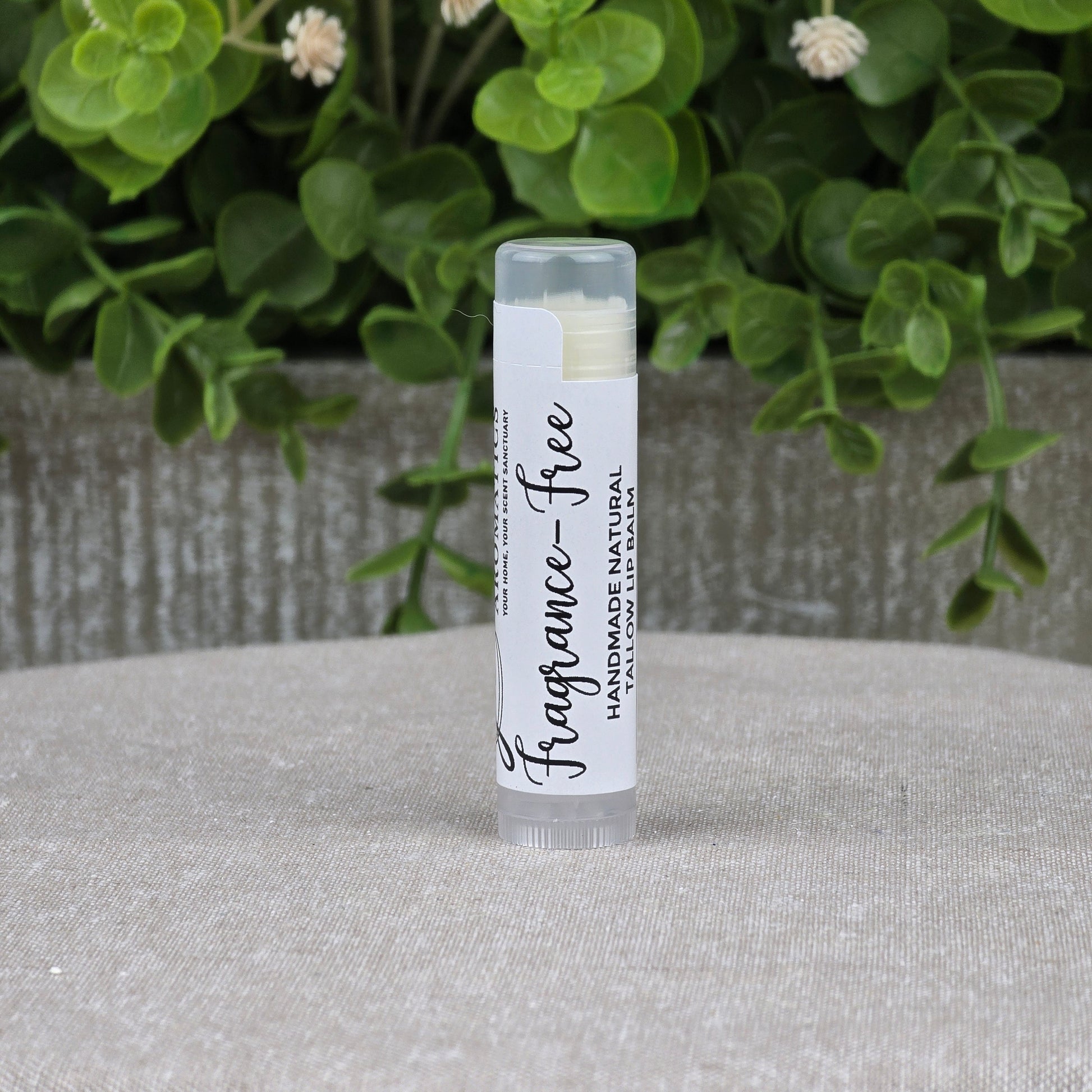 A tube of Jarhead Aromatics Tallow Handmade Natural Lip Balm – Assorted Flavors stands upright on a gray surface, with green leafy plants blurred in the background. This nourishing lip balm features pure ingredients and is handmade for healthy lips.
