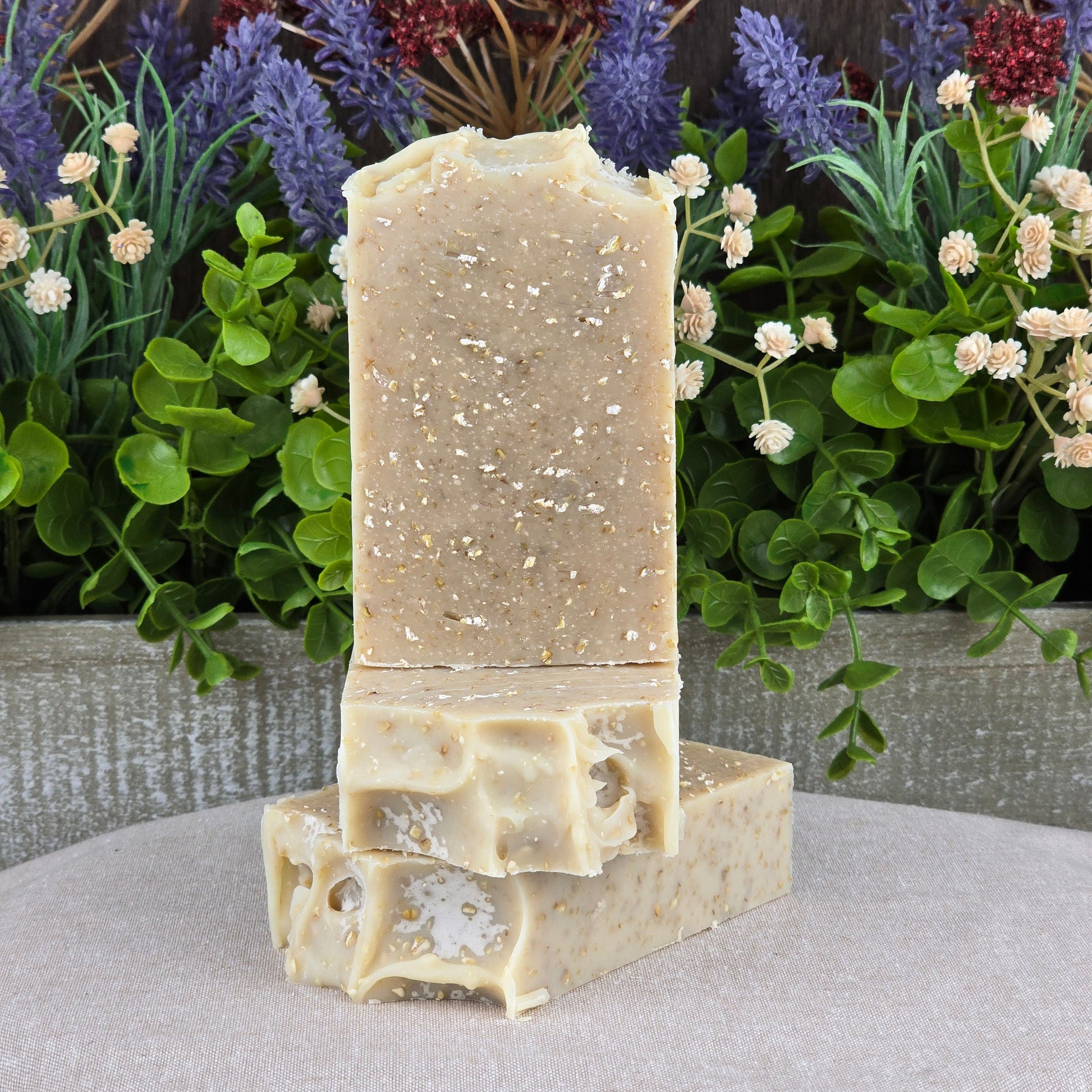 Two beige Fragrance-Free Oatmeal Handmade Artisan Soaps by Jarhead Aromatics, featuring visible oat flakes, are stacked on a neutral cloth amid green leaves and flowers—ideal for sensitive skin care and made with all-natural ingredients.