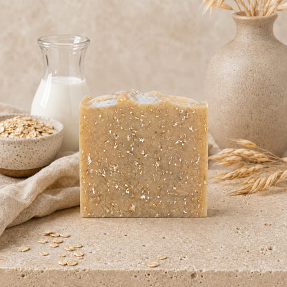 A Fragrance-Free Oatmeal Handmade Natural Goat Milk Artisan Soap bar by Jarhead Aromatics stands on a stone surface, accompanied by oats, a small jar of milk, a beige cloth, and a beige vase with dried grasses in the background, creating a natural, rustic scene. This Unscented Goat Milk & Oatmeal Soap is thoughtfully crafted for those who value simplicity and gentle care in their daily routine.