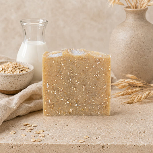 A Fragrance-Free Oatmeal Handmade Natural Goat Milk Artisan Soap bar by Jarhead Aromatics stands on a stone surface, accompanied by oats, a small jar of milk, a beige cloth, and a beige vase with dried grasses in the background, creating a natural, rustic scene. This Unscented Goat Milk & Oatmeal Soap is thoughtfully crafted for those who value simplicity and gentle care in their daily routine.