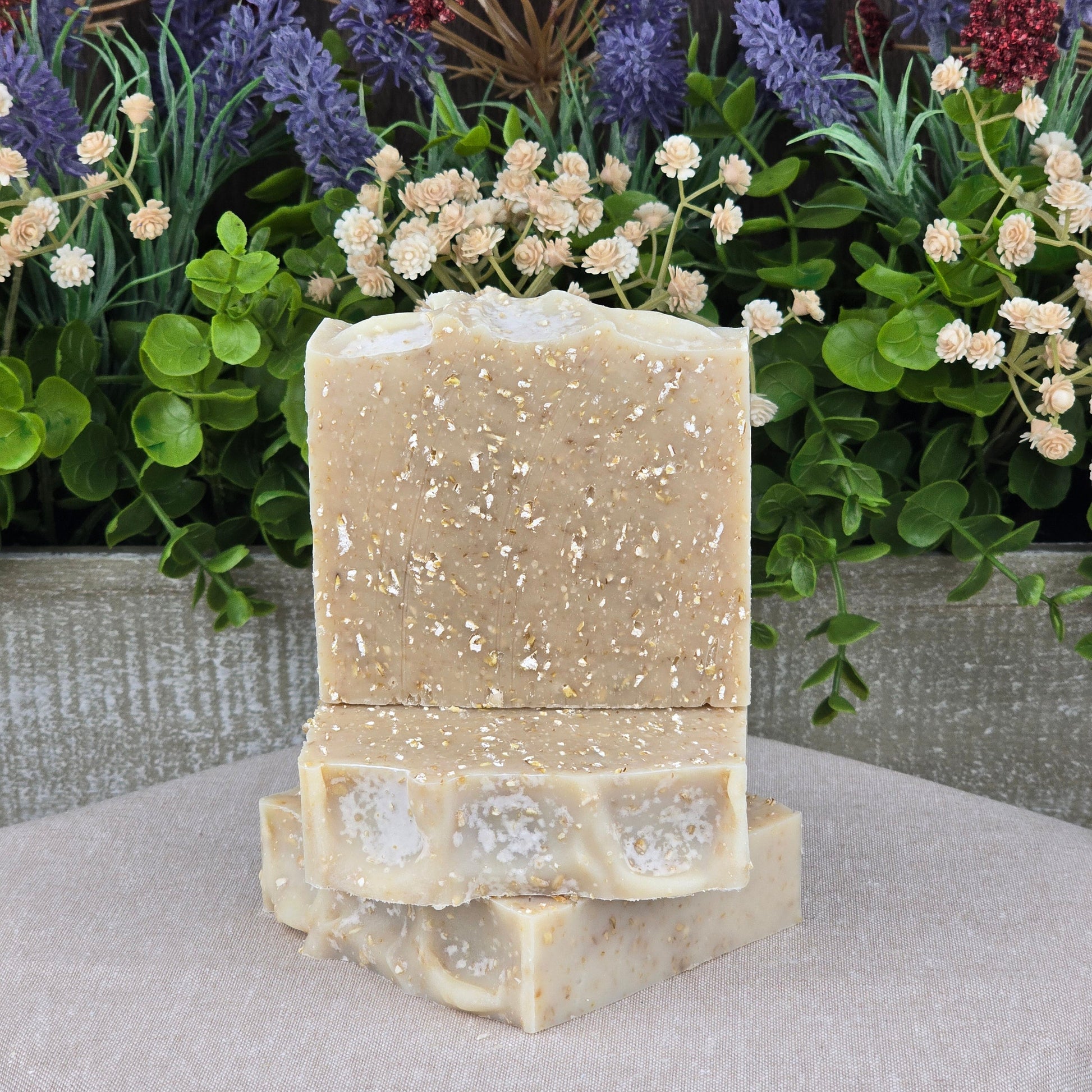 Two bars of Jarhead Aromatics Fragrance-Free Oatmeal Handmade Natural Goat Milk Artisan Soap, with a speckled texture, are stacked on a gray cloth. Artificial greenery and colorful flowers decorate the background—ideal for sensitive skin.