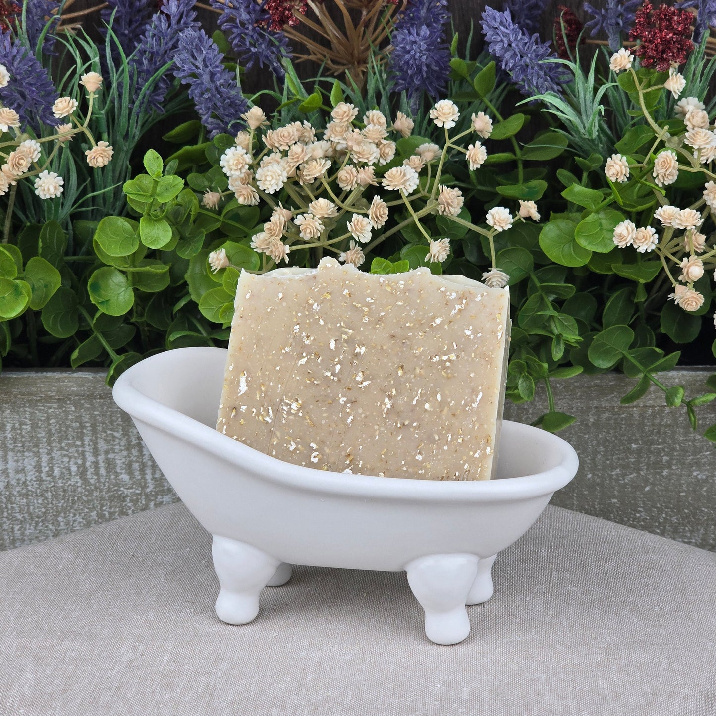 A bar of Jarhead Aromatics Fragrance-Free Oatmeal Handmade Natural Goat Milk Artisan Soap sits in a white ceramic bathtub-shaped dish, with artificial green and purple foliage and red flowers in the background.