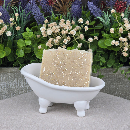 A bar of Jarhead Aromatics Fragrance-Free Oatmeal Handmade Natural Goat Milk Artisan Soap sits in a white ceramic bathtub-shaped dish, with artificial green and purple foliage and red flowers in the background.