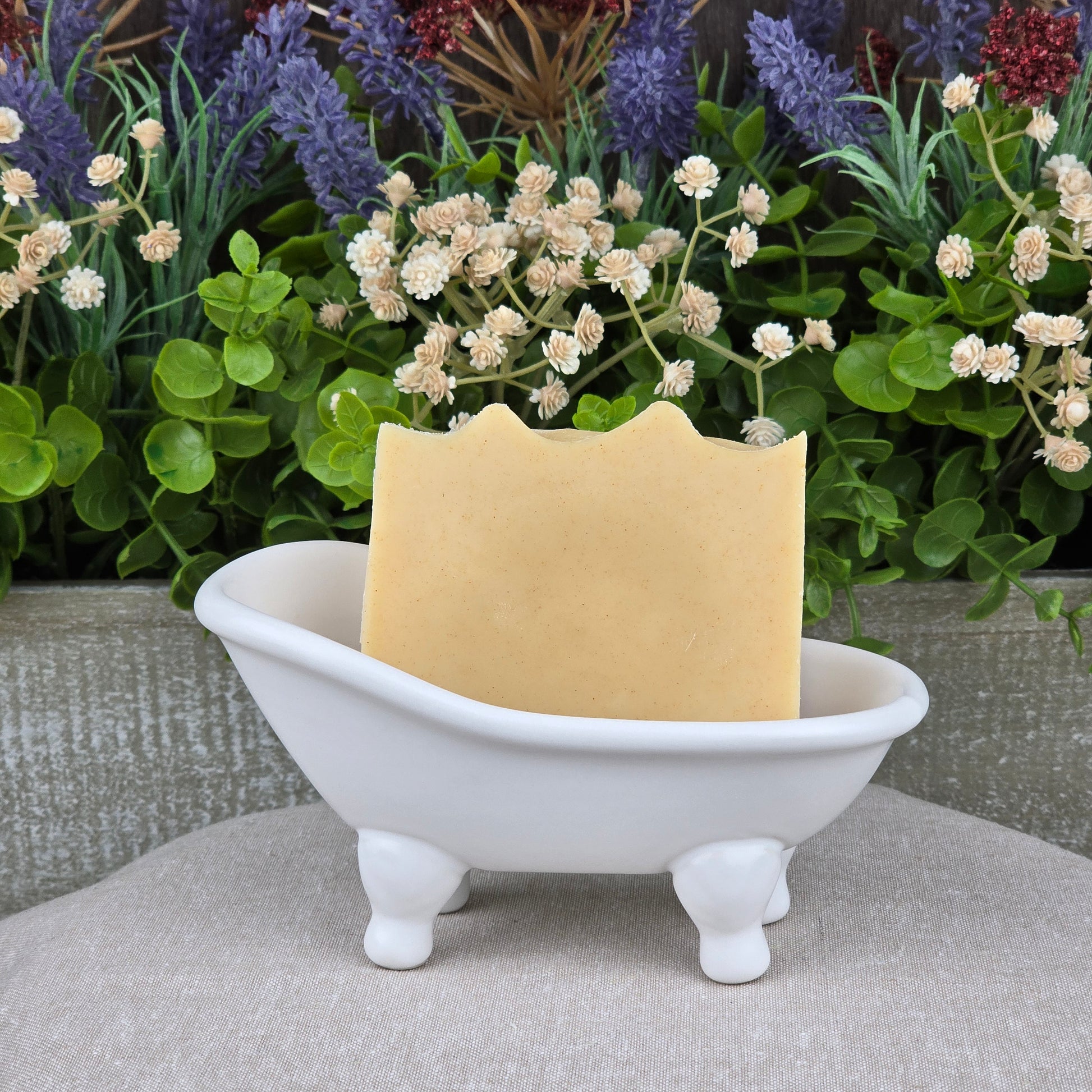 A bar of Jarhead Aromatics Frankincense + Citrus Handmade Natural Tallow Artisan Soap rests in a white clawfoot bathtub-shaped dish, with colorful flowers and green plants in the background.