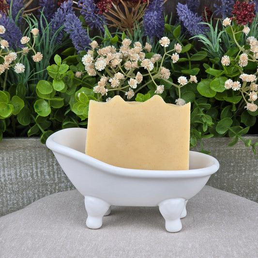 A bar of Jarhead Aromatics Frankincense + Citrus Handmade Natural Tallow Artisan Soap rests in a white clawfoot bathtub-shaped dish, with colorful flowers and green plants in the background.