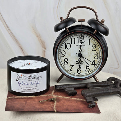 A black alarm clock reads 8:28 beside Jarhead Aromatics Galactic Twilight Coconut Wax Blend Candle from the Timeless Collection, a brown envelope topped with a gold key, and two large vintage keys on a marble-patterned light surface.