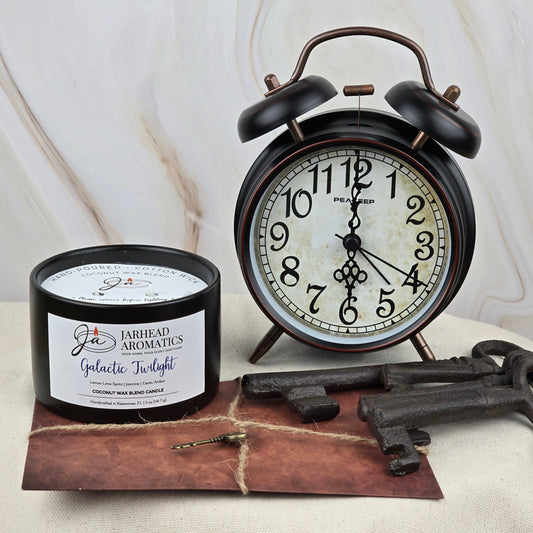A black alarm clock reads 8:28 beside Jarhead Aromatics Galactic Twilight Coconut Wax Blend Candle from the Timeless Collection, a brown envelope topped with a gold key, and two large vintage keys on a marble-patterned light surface.